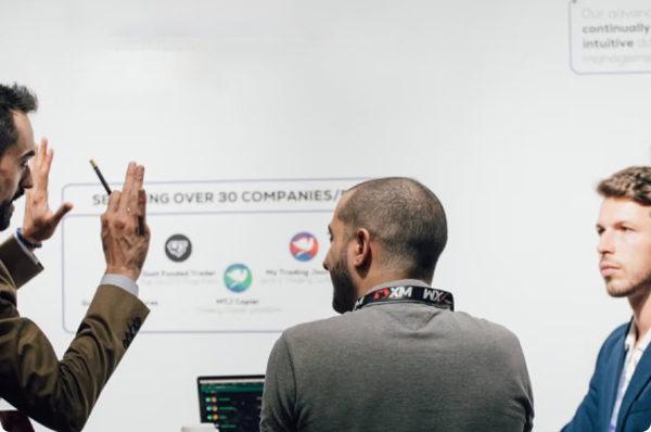 Man with a pen explaining to a group in front of a Serving over 30 companies board at an expo booth