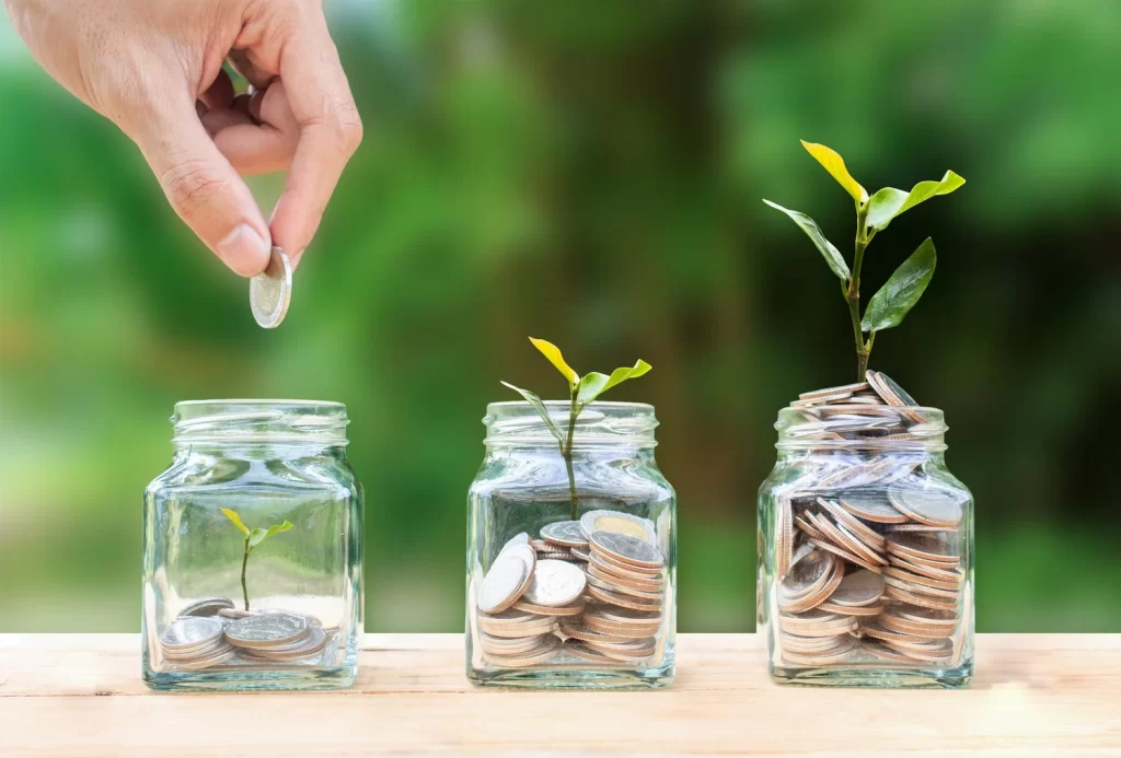 Hand dropping a coin into glass jars of coins with young plants growing, symbolizing savings