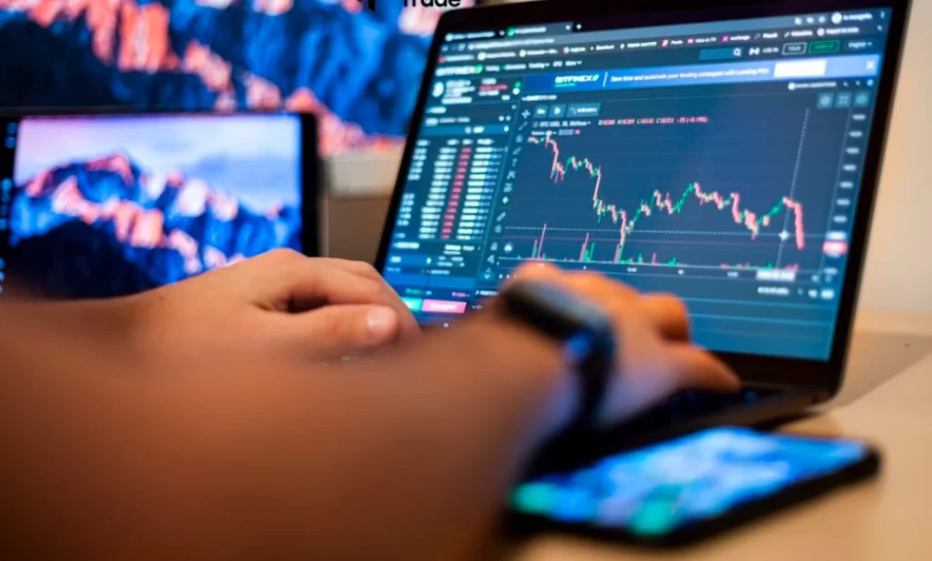 Hands of a trader using a laptop with candlestick charts and a smartphone on a desk