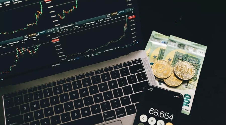 Laptop with trading charts on a dark desk beside banknotes, coins and a calculator
