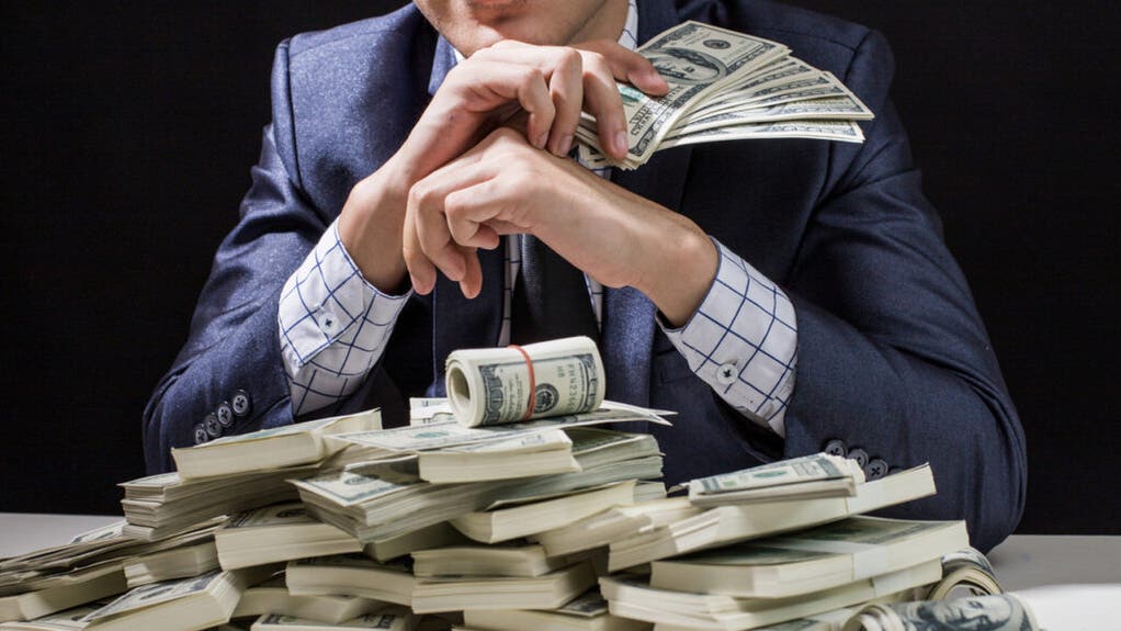 Businessman in a suit sitting behind stacks of dollar bills, hands clasped around cash