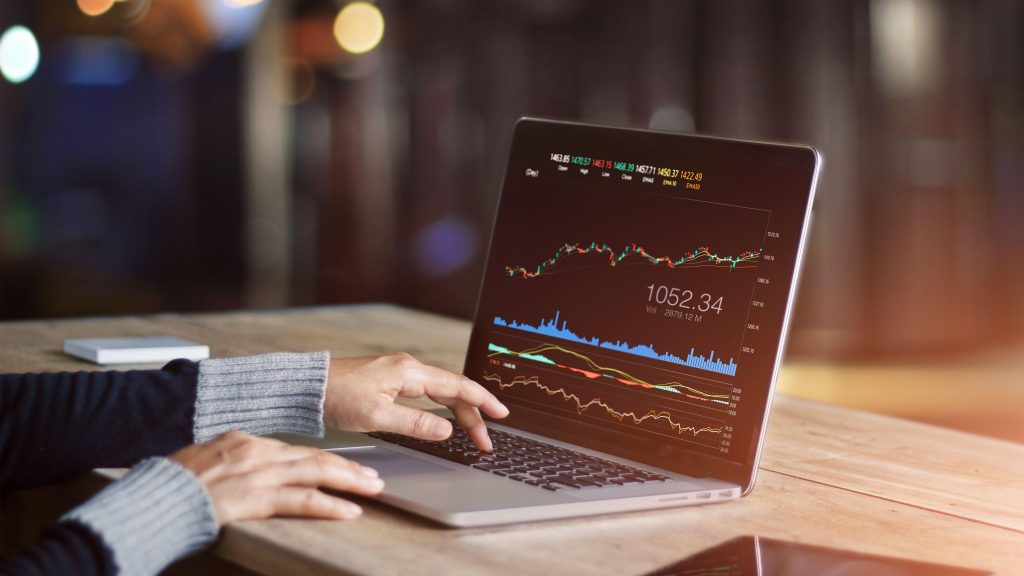 Hands typing on a laptop that shows trading charts, on a wooden table in warm light