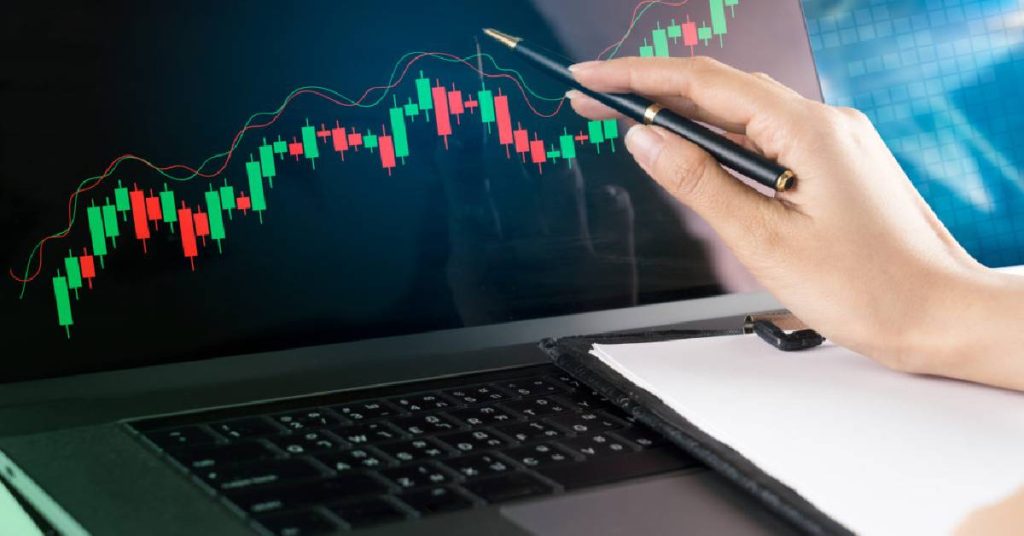 Hand pointing a pen at a rising candlestick chart on a laptop screen during analysis