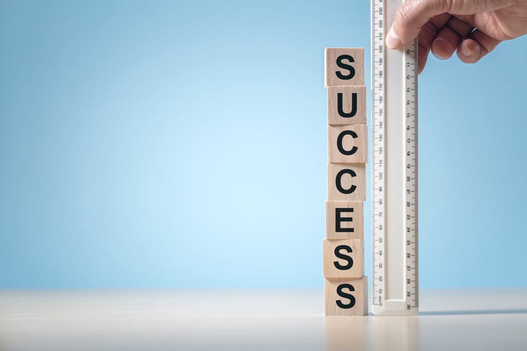 Wooden blocks spelling “SUCCESS” being measured with a ruler against a light blue background